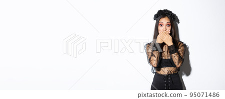 Shocked woman in witch outfit gasping and cover mouth speechless, standing over white background, looking at something surprising, wearing black lace dress and wreath, white background 95071486
