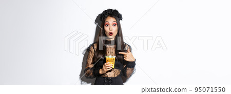 Amazed asian girl looking at camera while pointing finger at sweets, gather treats on halloween, wearing witch dress, standing over white background 95071550