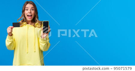 Amused and excited blond girl recommend using banking service, showing smartphone display and credit card and looking camera fascinated as receive bonus to bank account, blue background 95071579
