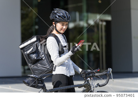 一個送餐的女人,手裡拿著智能手機,拉著一輛公路自行車 一個送餐的女人,手裡拿著智能手機,拉著一輛公路自行車 95073401