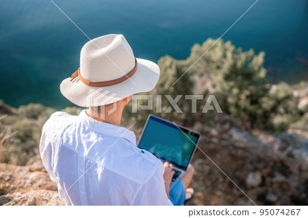 Digital nomad, man in the hat, a businessman with a laptop sits on the rocks by the sea during sunset, makes a business transaction online from a distance. Remote work on vacation. Digital nomad, man in the hat, a businessman with a laptop sits on the rocks by the sea during sunset, makes a business transaction online from a distance. Remote work on vacation. 95074267