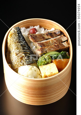 A grilled fish bento box containing pork, potato salad, omelet, etc. is photographed against a black background. 95074954