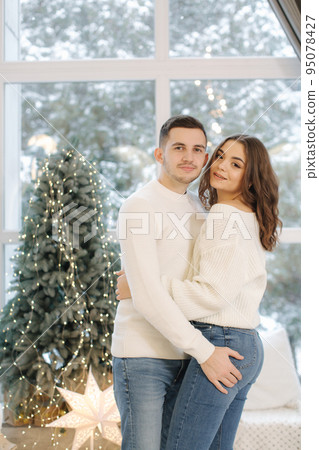 Beautiful young couple posing to photographer at New Year studio. Christmas tree. Snow outside Beautiful young couple posing to photographer at New Year studio. Christmas tree. Snow outside 95078427