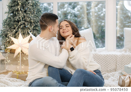 Handsome boy with his beautiful girlfriend hug each other at home at Christmas eve. Fit tree and snow outsif. Christmas mood 95078450