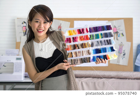 Fashion designer room. Young asian dressmaker present silk thread color sample catalog with a smile. 95078740
