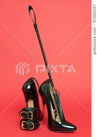 Adult sex games. Kinky lifestyle. Spank and a pair of black high-heeled shoes on the red background. Bdsm outfit 95080287
