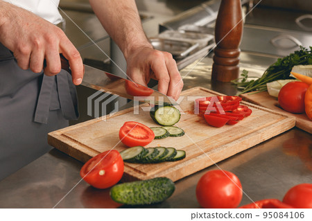 Busy chef cutting tomatoes and cucumbers on a board for making salad in modern restaurant kitchen 95084106