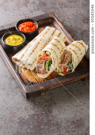Doner kebab made of meat cooked on a vertical rotisserie with additions of vegetables and sauce wrapped in lavash closeup on the wooden board. Vertical 95085046