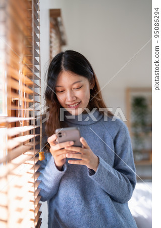 Cheerful young asian woman using mobile phone near window at home. 95086294