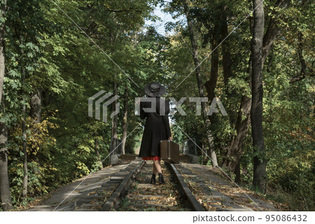 Woman in black coat and hat of 40s style with retro valise in hands walks along rails going through forest. Back view Woman in black coat and hat of 40s style with retro valise in hands walks along rails going through forest. Back view 95086432