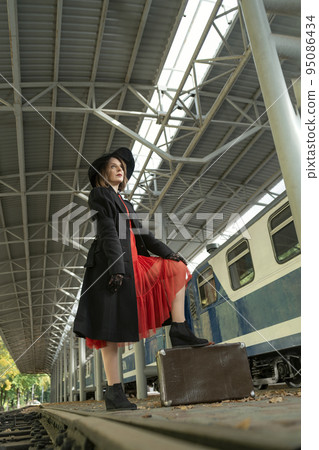 Young woman on platform near train with leather suitcase wearing in red dress, black coat and hat . Vertical frame Young woman on platform near train with leather suitcase wearing in red dress, black coat and hat . Vertical frame 95086434