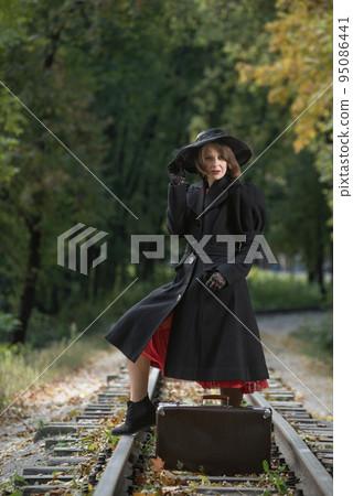 Middle-aged woman with old suitcase in red dress black hat and coat poses on rails on wood background. Vertical frame Middle-aged woman with old suitcase in red dress black hat and coat poses on rails on wood background. Vertical frame 95086441
