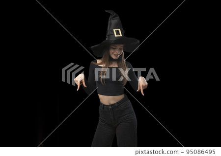 Girl in black cloth and hat points fingers down. Teenager in image of witch on black background. Copy space Girl in black cloth and hat points fingers down. Teenager in image of witch on black background. Copy space 95086495