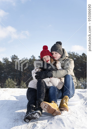Young guy and a girl sit and hug on the snow against the background of the forest and the blue sky. Vertical frame Young guy and a girl sit and hug on the snow against the background of the forest and the blue sky. Vertical frame 95086690
