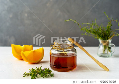 Thyme honey in small jar, wooden tea spoon, small thyme branch, orange slices on white table with grey background 95091056