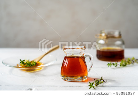 Thyme honey in small jug and jar, small thyme branch on white wooden table with grey background 95091057