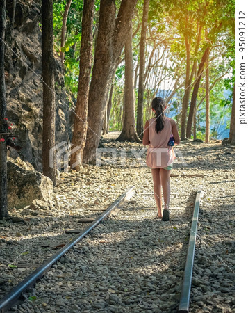 Back view of young girl with water bottle walking alone in forest nature path walk on trail woods background at Hellfire Pass in Kanchanaburi Thailand. Happy girl relaxing on active outdoor activity. 95091212