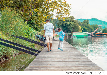 Back view of Asian grandfather and grandchild wear protective face mask to prevent Coronavirus (COVID-19) walking in a nature path on wooden bridge along river. Together outdoors family concept. 95091213
