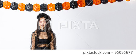 Portrait of elegant asian woman in witch costume, looking confident and standing against pumpkin streamers, decorations for halloween over white background Portrait of elegant asian woman in witch costume, looking confident and standing against pumpkin streamers, decorations for halloween over white background 95095677
