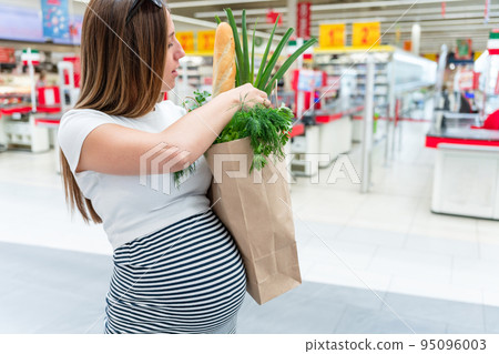 Woman supermarket grocery food bag. Pregnancy mother with healthy vegetables, fresh lettuce salad leaves in market food bag on grocery supermarket background. Everyday shopping concept. 95096003