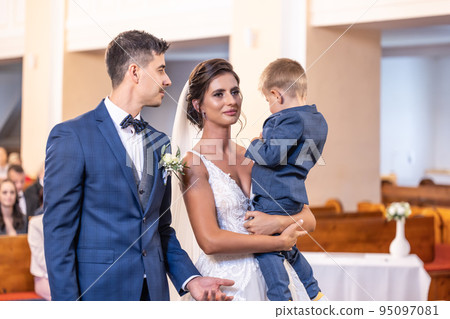Happy young newlyweds stand in the church with the bride holding a young son in her arms 95097081