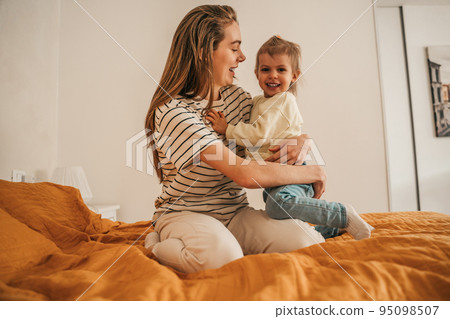 Joyous female parent embracing her child seated in the bedroom Joyous female parent embracing her child seated in the bedroom 95098507