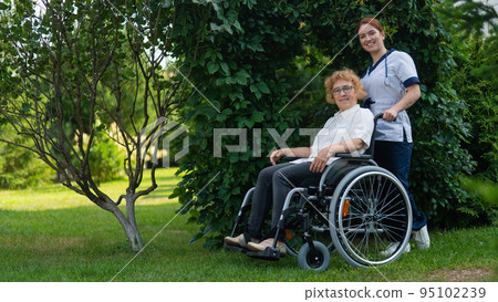 Caucasian female doctor walks with an elderly patient in a wheelchair in the park. Nurse accompanies an old woman on a walk outdoors.  95102239