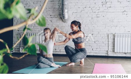 Pretty blond female yoga student is learning arm twists in cow face position from friendly cheerful instructor on personal training. Women are talking, smiling and enjoying practice. Pretty blond female yoga student is learning arm twists in cow face position from friendly cheerful instructor on personal training. Women are talking, smiling and enjoying practice. 95106144