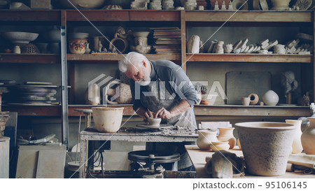 Experienced male ceramist is creating utensils from clay on potter's wheel in workshop. Producing eathenware, handmade utensils and professional pottery concept. Experienced male ceramist is creating utensils from clay on potter's wheel in workshop. Producing eathenware, handmade utensils and professional pottery concept. 95106145