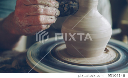 Elderly bearded craftsman is creating ceramic jar on potter's wheel using professional tools. Workplace with handmade pots, beautiful vases and figures in background. Elderly bearded craftsman is creating ceramic jar on potter's wheel using professional tools. Workplace with handmade pots, beautiful vases and figures in background. 95106146