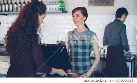 Cheerful workers of cozy coffeehouse are greeting customers, taking payments with smartphone using bank cards terminal and selling takeaway drinks to young people. 95106181