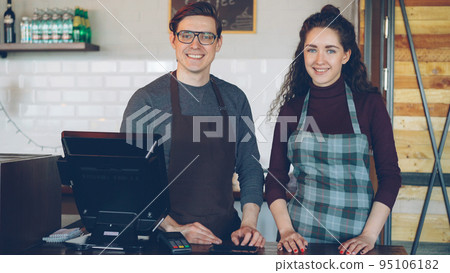 Portrait of two young good-looking waiters standing at cashier's desk in modern coffee-house and smiling. Successful business, happy people and food service concept. 95106182
