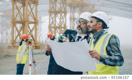 Architect man in suit is talking to engineer in building area while surveyors are working with modern equipment in background. People and business concept. Architect man in suit is talking to engineer in building area while surveyors are working with modern equipment in background. People and business concept. 95106187