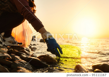 Coastal cleanup and garbage collection for recycling. A woman volunteer collects plastic bottle by the sea or river, closeup of hand. Low angle view. World environment day and Earth day concept 95111167