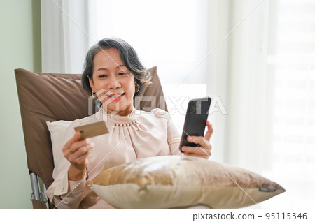 Senior Asian female using mobile banking application while relaxing in her living room. Senior Asian female using mobile banking application while relaxing in her living room. 95115346