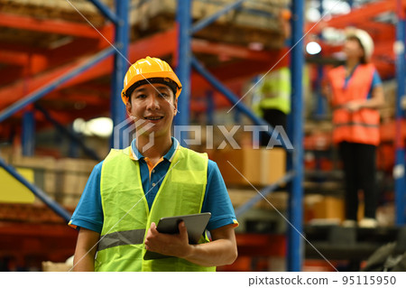 Portrait of male warehouse worker wearing safety hardhat and vest holding digital tablet standing in large retail warehouse 95115950