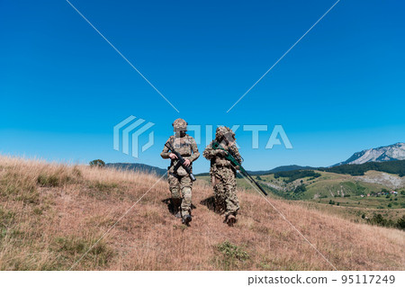 A sniper team squad of soldiers is going undercover. Sniper assistant and team leader walking and aiming in nature with yellow grass and blue sky. Tactical camouflage uniform. A sniper team squad of soldiers is going undercover. Sniper assistant and team leader walking and aiming in nature with yellow grass and blue sky. Tactical camouflage uniform. 95117249