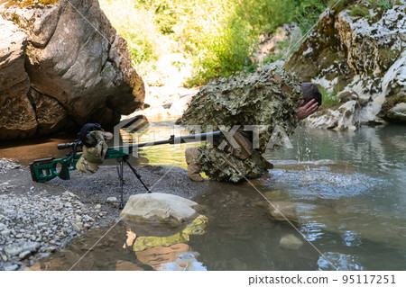 Soldier in a camouflage suit uniform drinking fresh water from the river. Military sniper rifle on the side. Soldier in a camouflage suit uniform drinking fresh water from the river. Military sniper rifle on the side. 95117251