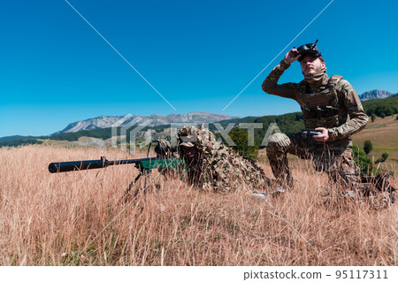 Sniper soldier assisted by an assistant to observe the area to be targeted with modern warfare tactical virtual reality goggles aerial drone military technology. 95117311
