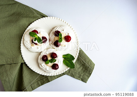 Meringue cakes decorated with whipped cream, cherries and mint leaves on a white plate and green napkin, top view with copy space Meringue cakes decorated with whipped cream, cherries and mint leaves on a white plate and green napkin, top view with copy space 95119088