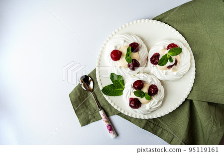 Meringue cakes decorated with whipped cream, cherries and mint leaves on a white plate and green napkin, top view with copy space Meringue cakes decorated with whipped cream, cherries and mint leaves on a white plate and green napkin, top view with copy space 95119161