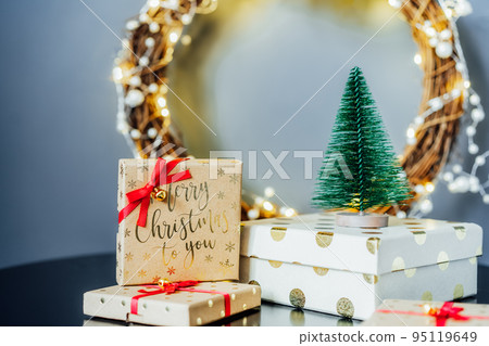 Christmas composition with gift boxes, red lantern, small Christmas trees decor and elegant flower wreath on black table on gray wall background. Festive home decor. Scandinavian style. Copy space Christmas composition with gift boxes, red lantern, small Christmas trees decor and elegant flower wreath on black table on gray wall background. Festive home decor. Scandinavian style. Copy space 95119649