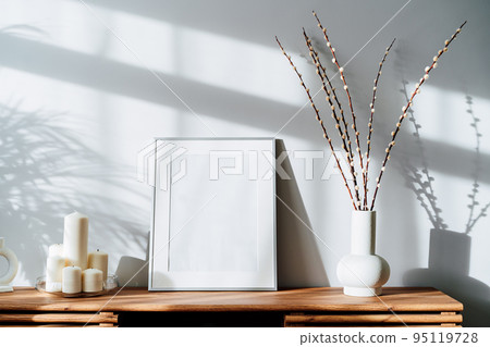 Modern minimalist style interior with white poster mockup, candles and blooming branches of the pussy willow in vase on wooden console under sunlight and shadows on a white gray wall. Selective focus 95119728