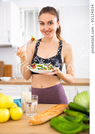 Portrait of woman standing in home kitchen and tasting vegetable salad Portrait of woman standing in home kitchen and tasting vegetable salad 95124834