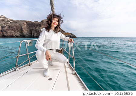 A woman sits on the bow of a yacht on a sunny summer day, the breeze develops her hair, a beautiful sea is in the background 95126626