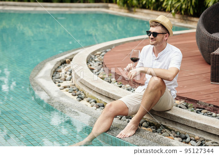 Young man enjoying glass of red wine while resting near the poolside outdoors Young man enjoying glass of red wine while resting near the poolside outdoors 95127344