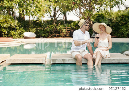Young couple toasting with glasses of red wine and celebrating their summer vacation while sitting at outdoor pool 95127462