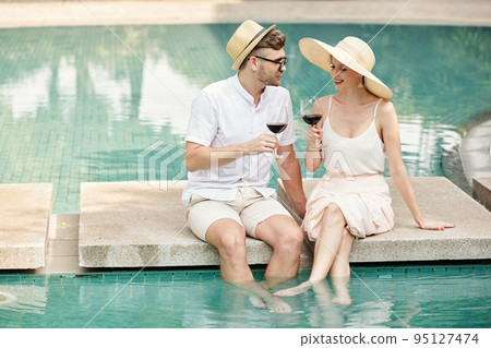 Young couple drinking red wine and talking sitting at outdoor pool at resort during vacation 95127474