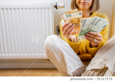 Warmly dressed woman counting money near radiator 95127578