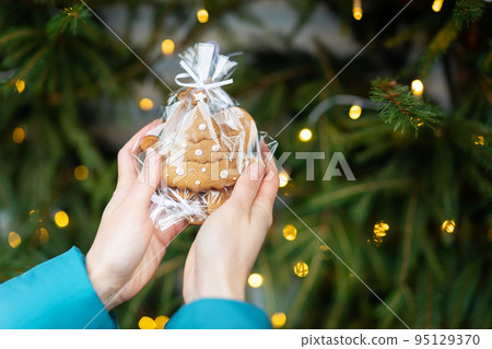 A woman holds in her hands gingerbread in the form of a Christmas tree on the background of a green Christmas tree outdoors. The concept of winter holidays, sweets and coziness. 95129370